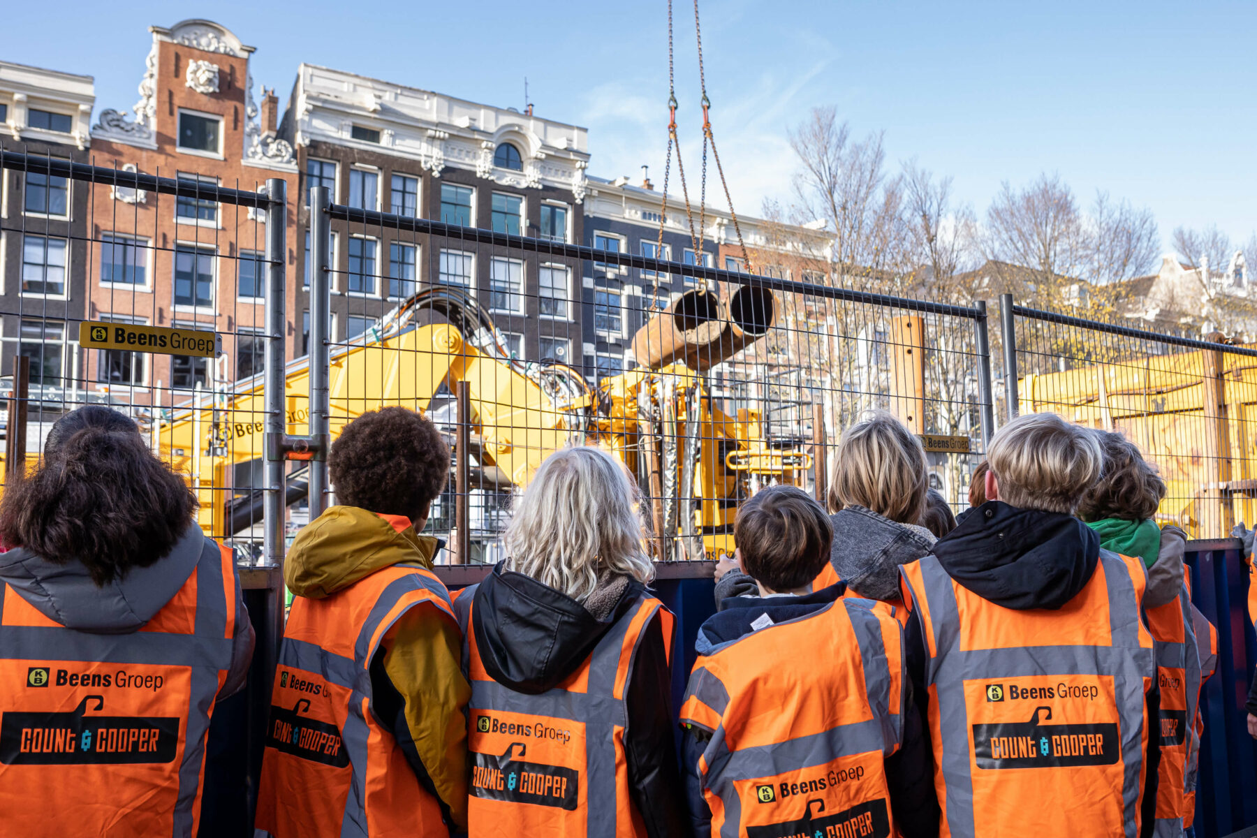 Een groep leerlingen in oranje veiligheidshesjes kijkt door de bouwhekken heen naar de werkzaamheden aan de kademuur.