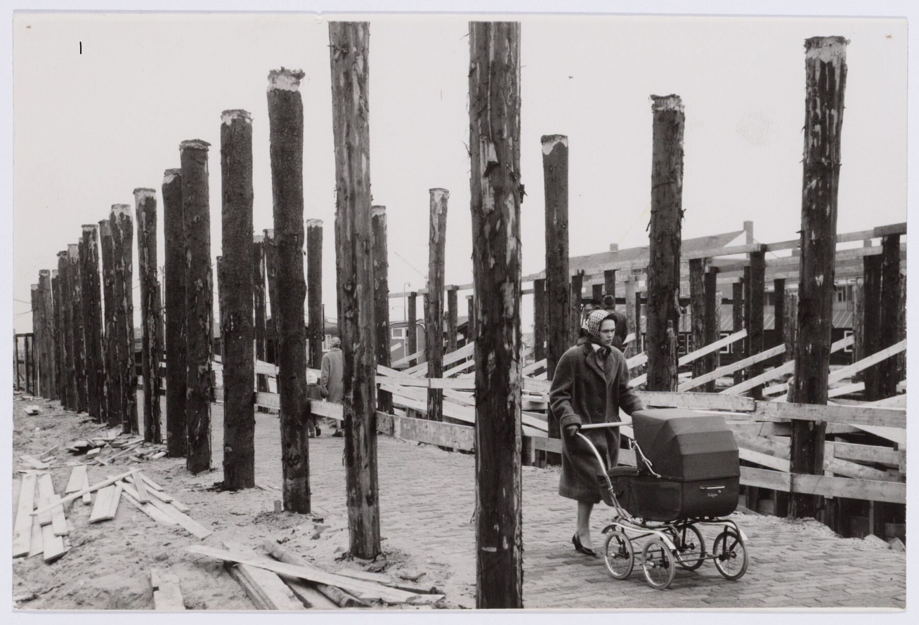 Vrouw met kinderwagen loopt tussen rijen opstaande palen voor een brug.