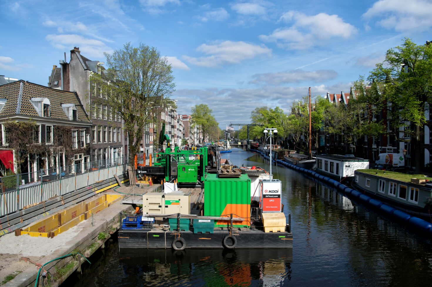 Bouwplaats van Koningsgracht met machines op ponton, sleufkist in de kade en woonboten aan de andere kant achter een ballenlijn. De vaarweg is open.