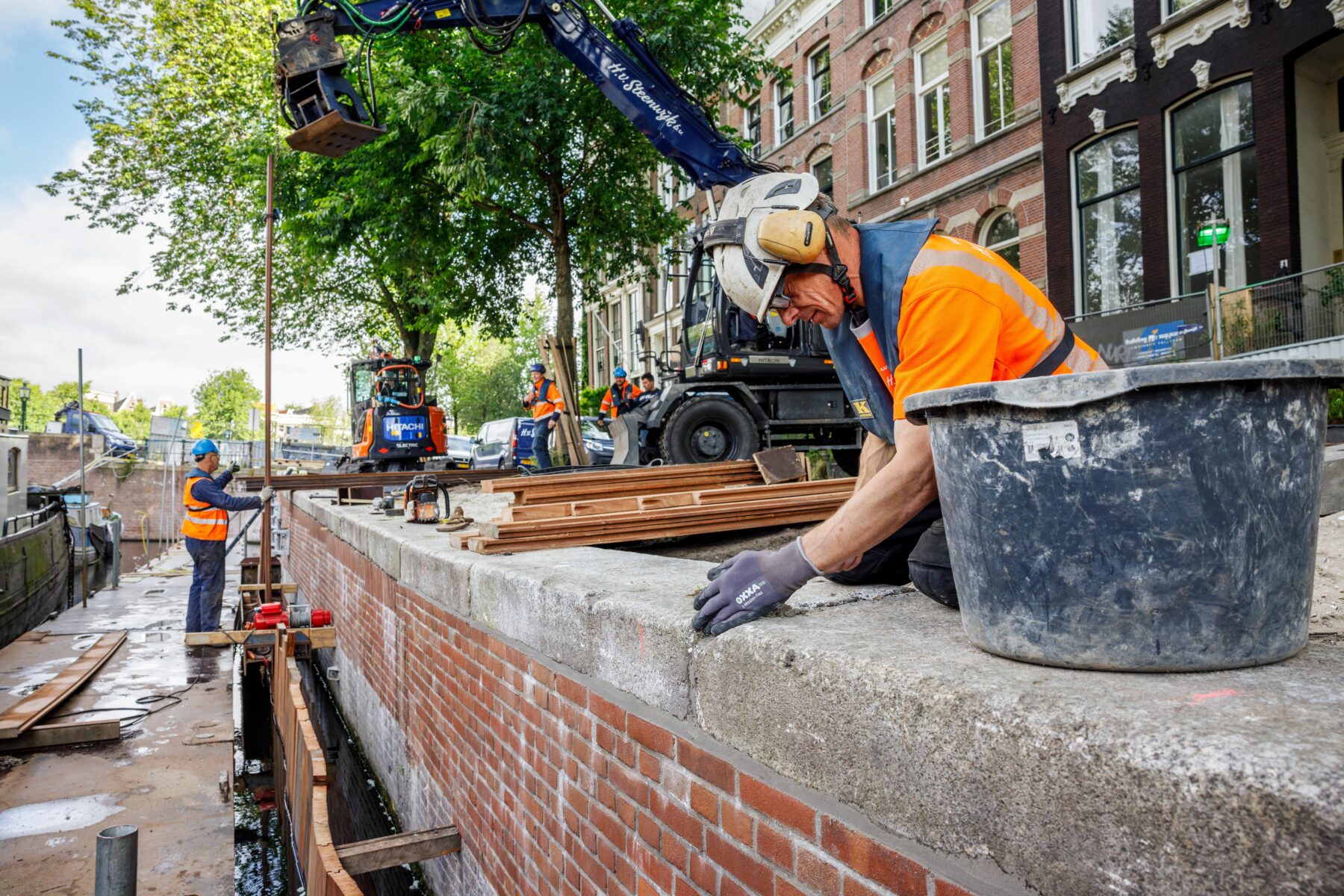 Man plaatst een originele deksteen terug op de kademuur. Op de achtergrond werken ze aan het plaatsen van het onderloopheidsscherm