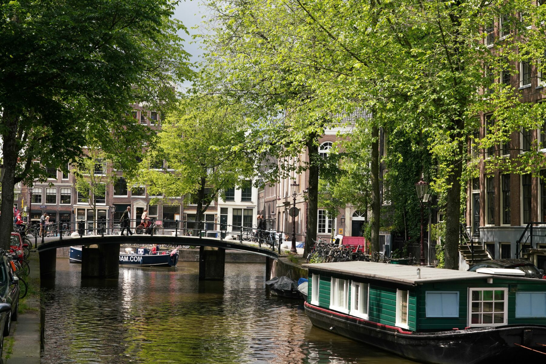 Idyllisch beeld van bomen langs de Raamgracht, met een woonboot op de gracht en een man die over een bruggetje loopt