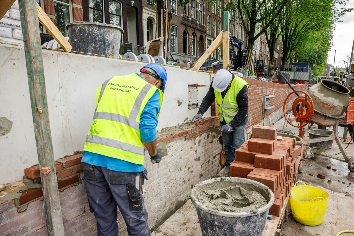 Twee metselaars metselen de voorkant van het nieuwe stuk kademuur tegen het nieuwe beton