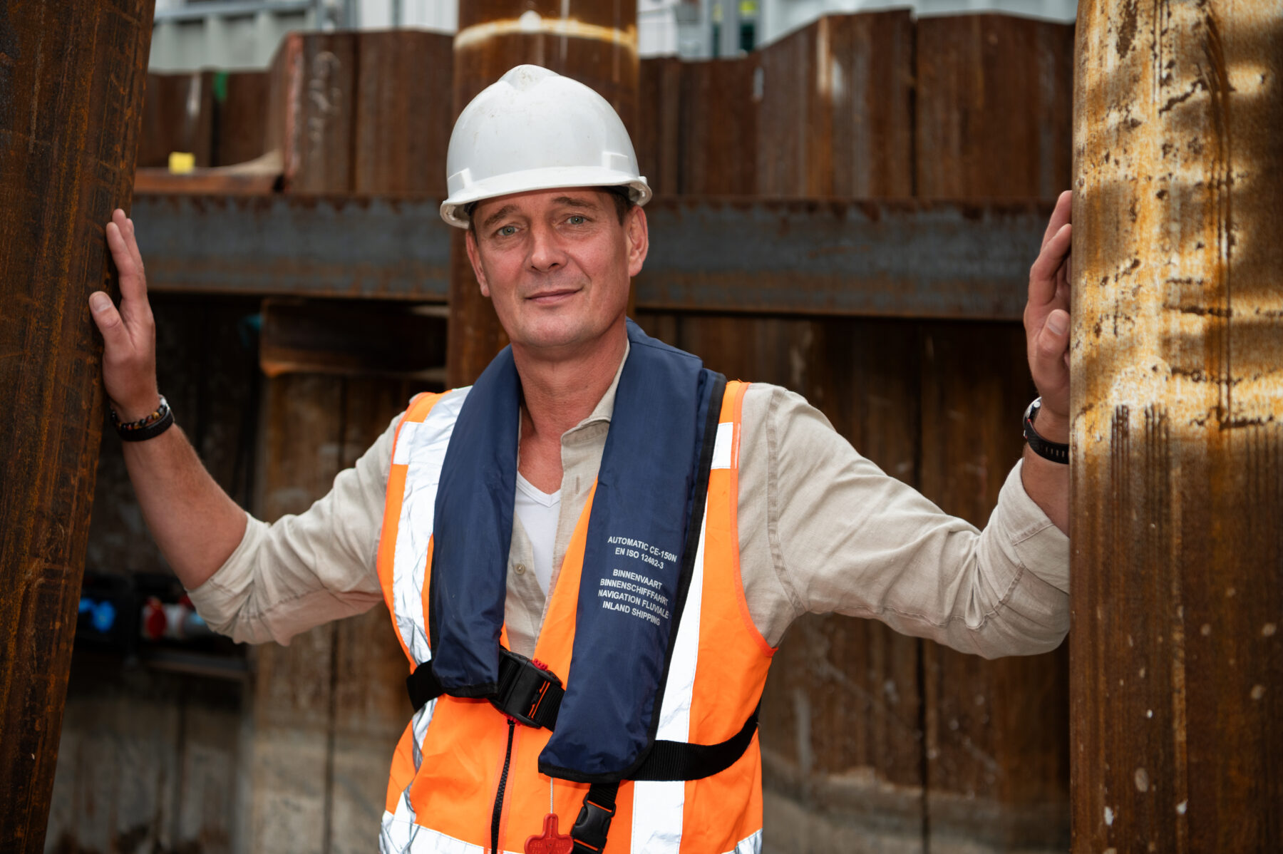 Directievoerder Jan Adriaanse poseert op de bouwplaats. Hij draagt de veiligheidskleding: een helm en een zwemvest.