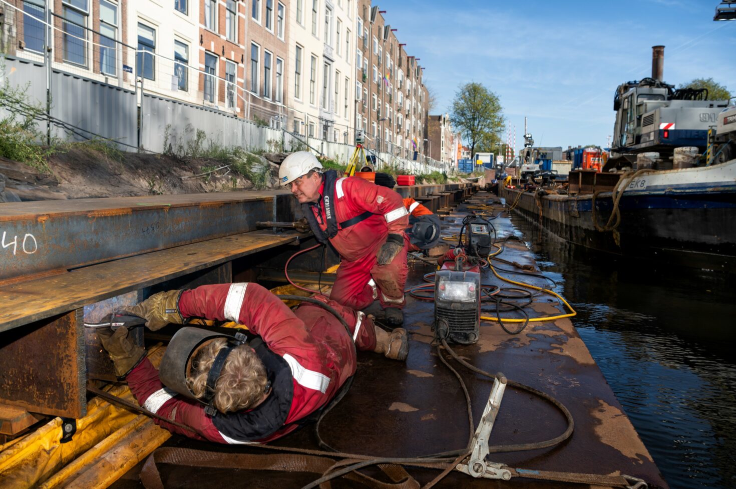 Zoon Rick Dobber maakt een las, liggend op een plankier voor de kade, terwijl vader Ron toekijkt