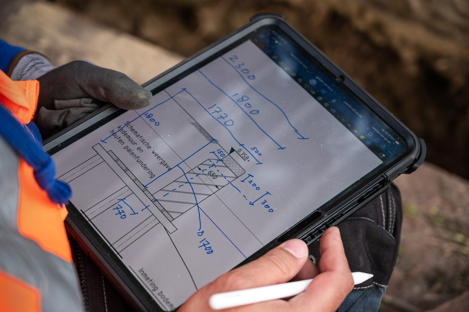en tablet met daarop een technische tekening van een kademuur. Hierop heeft Miguel de verschillende afmetingen genoteerd die ze zojuist hebben opgenomen, zoals de dikte van de kademuur en de lengte en diepte van de kadevloer.