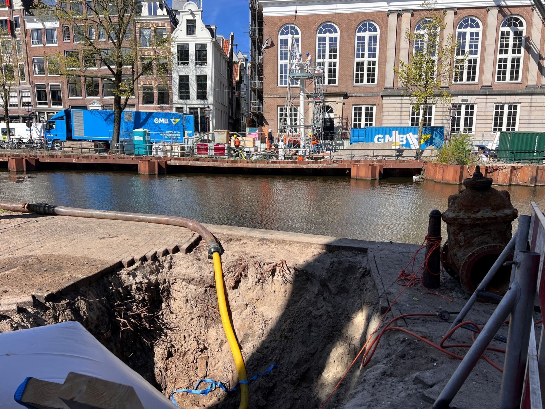Kloveniersburgwal met gat op de voorgrond en stellage aan de overkant van de gracht. De kous wordt zo door de bestaande drinkwaterleiding van de ene kant naar de andere kant van de gracht geschoven.