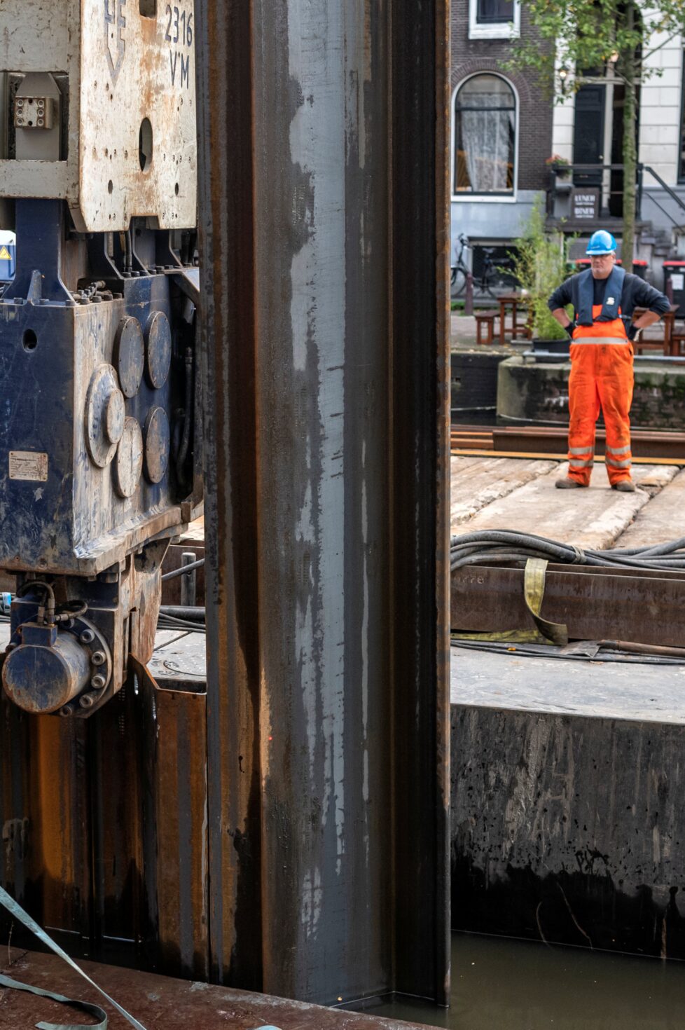 Versterking van kademuren. Werkman kijkt vanaf de kant naar een lange stalen damwandplank op de voorgrond