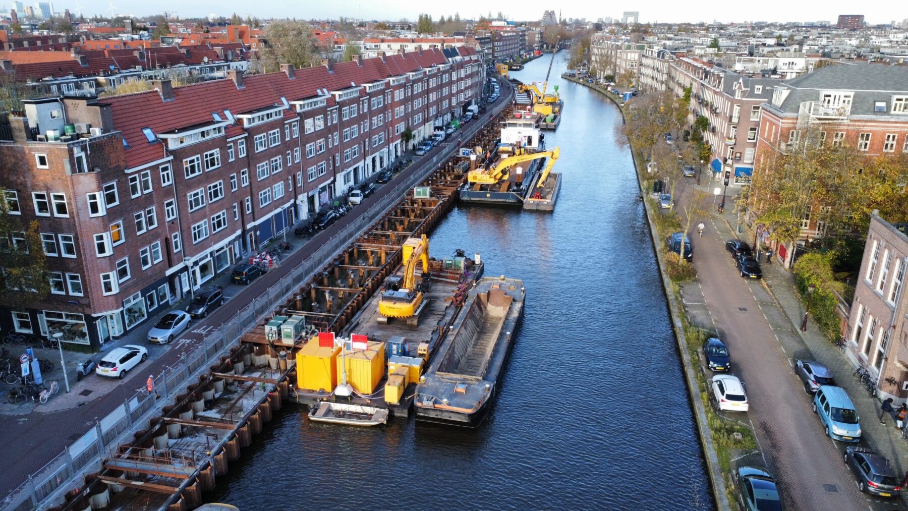 ogelperspectief op de Kostverlorenvaart met een open kade waar de kademuur wordt gemaakt en bulldozers op pontons op de gracht, daarnaast duwbakken voor aan- en afvoer van materialen. Op de achtergrond de Pontsteiger.