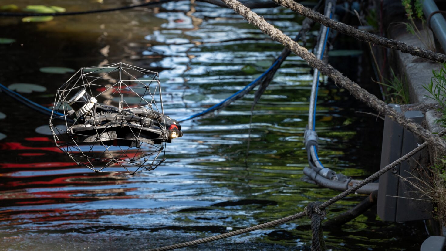 Een drone zweeft net boven het water bij een kademuur langs de Amstel