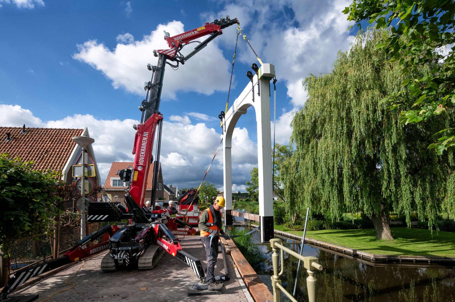 De hameipoort, het traditionele bovenwerk van een ophaalbrug, hangt aan een kleine kraan boven de Bakkersbrug om daar geinstalleerd te worden. Op de voorgrond en achtergrond werkers die de brug op z'n plek zetten.