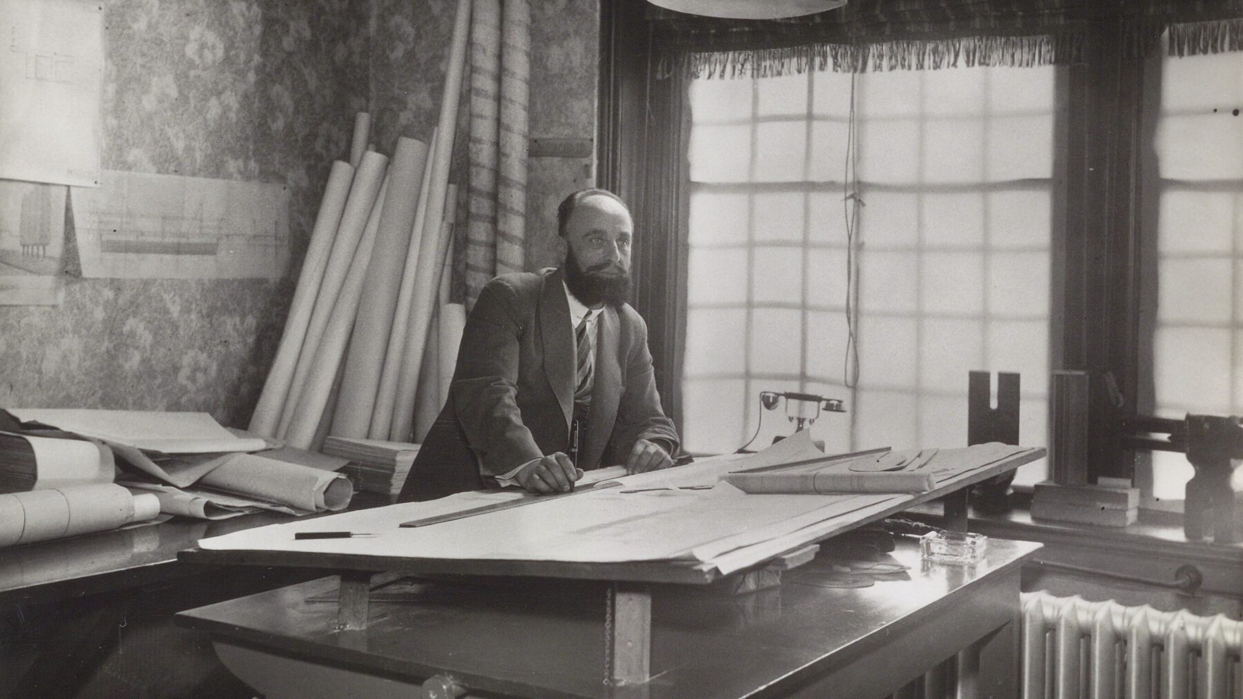 Piet Kramer poseert aan zijn tekentafel in het stadhuis, in 1938. Met op de achtergrond bouwtekeningen op de wand en rollen bouwtekeningen