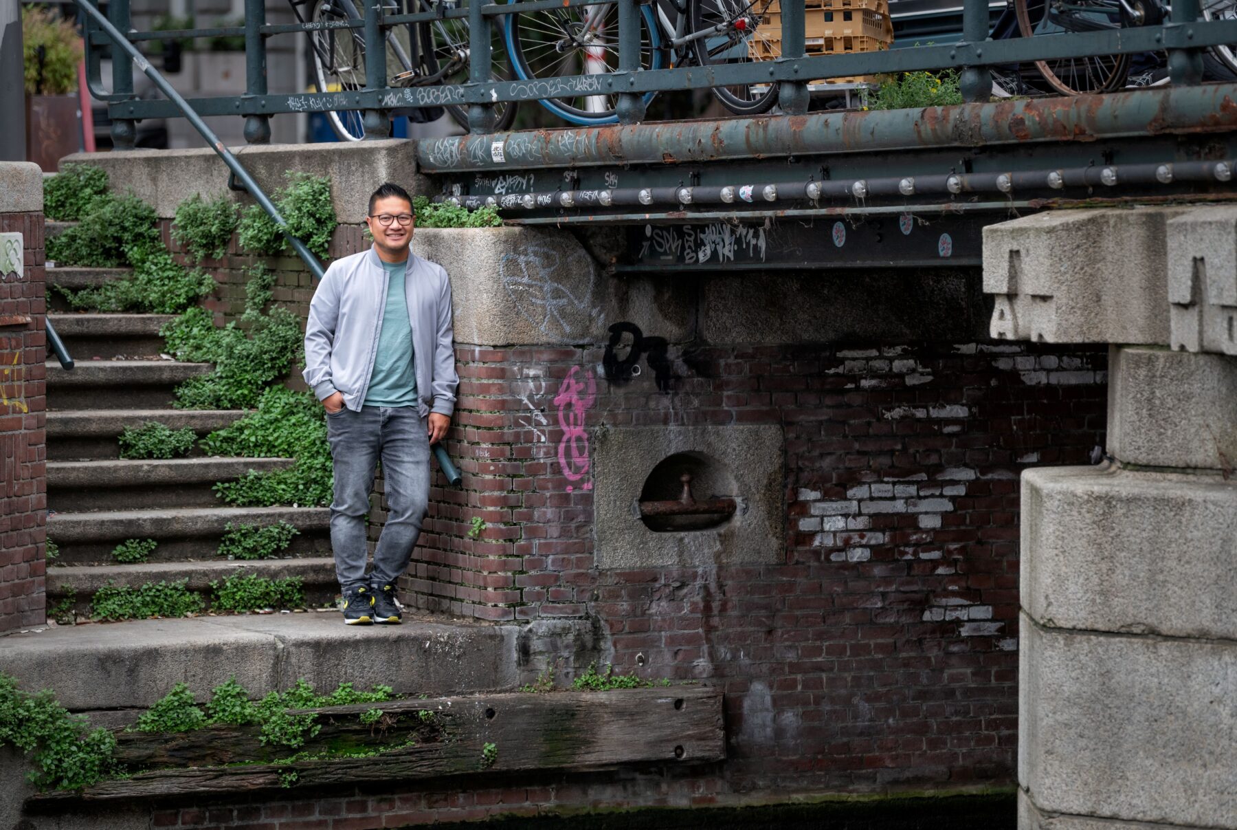 Constructeur Long Ha op een trapje naar de gracht bij de Doelensluis. Onder hem het water van het Rokin, boven hem wat fietsen aan de hekken op de brug